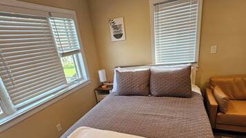 Un dormitorio con cama, dos ventanas con persianas, una mesita de noche, una lámpara, una obra de arte en la pared y un sillón de cuero marrón.