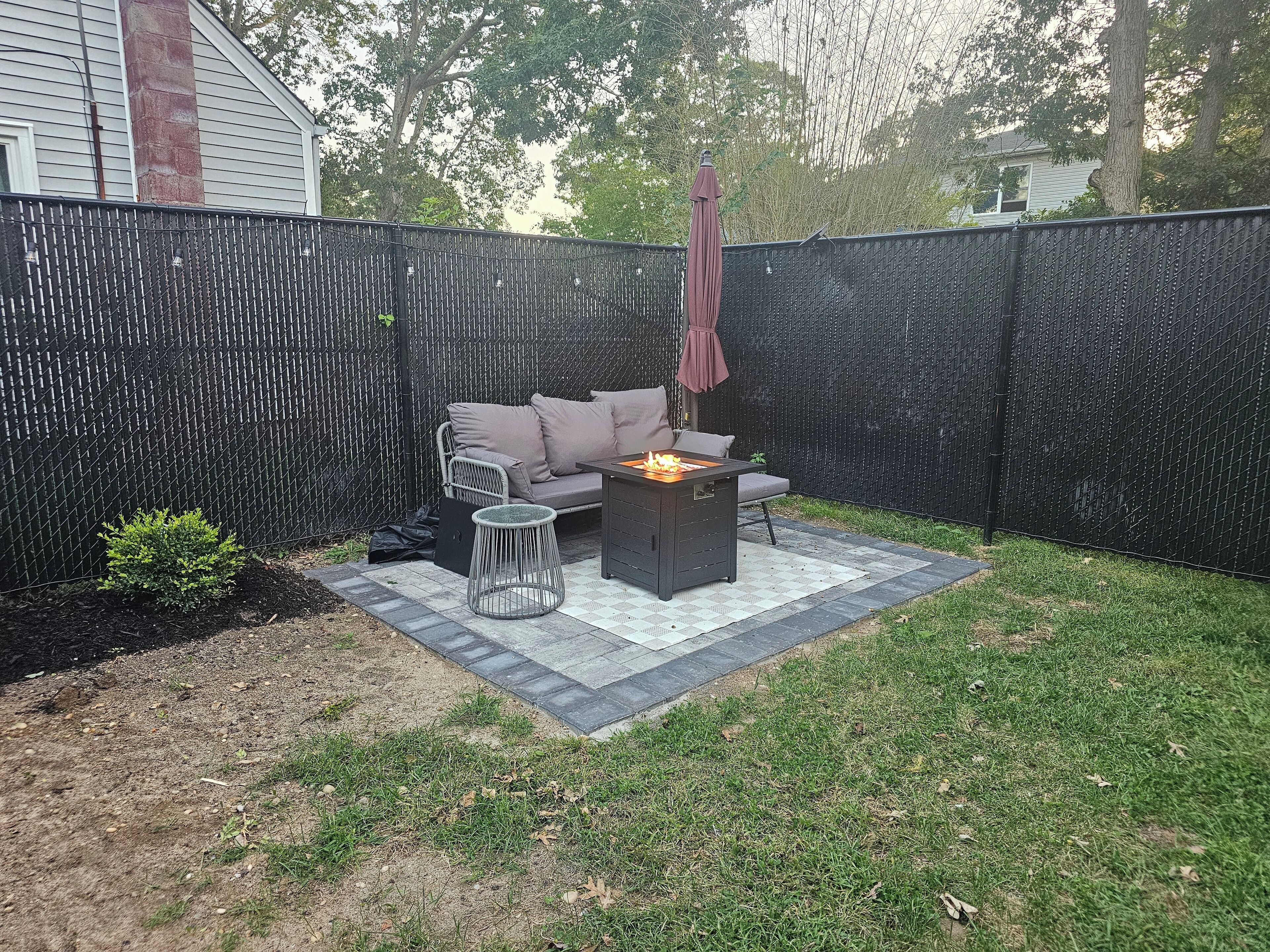 Un jardin avec un espace patio, une clôture en métal noir, un canapé gris, une table ronde et un foyer.
