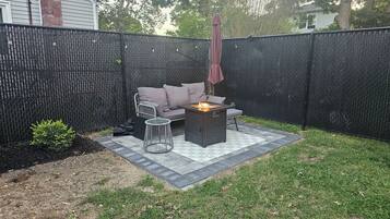 Un jardin avec un espace patio, une clôture en métal noir, un canapé gris, une table ronde et un foyer.