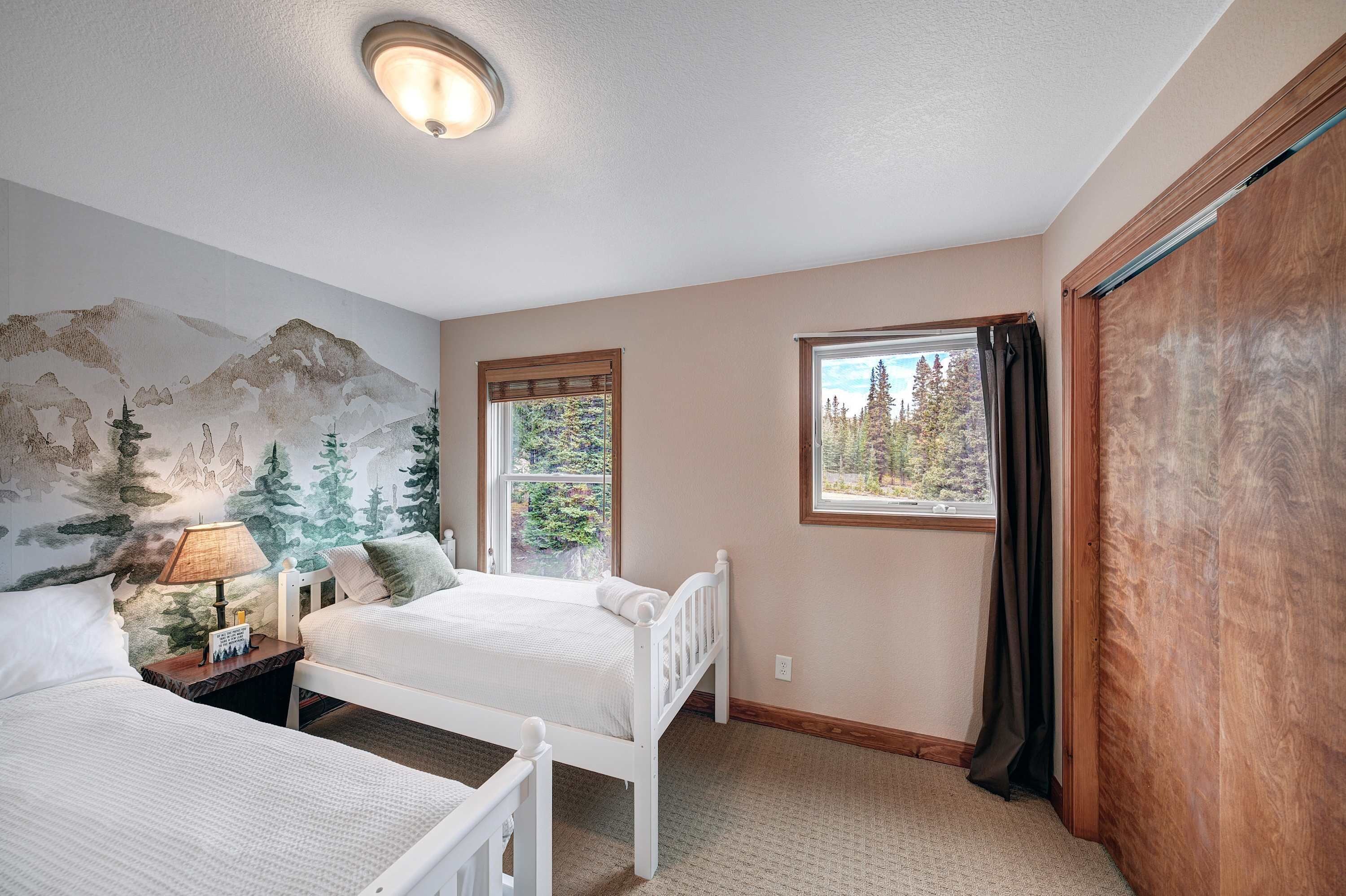 A bedroom with two beds, a nightstand with a lamp, a wall mural, and two windows with curtains.