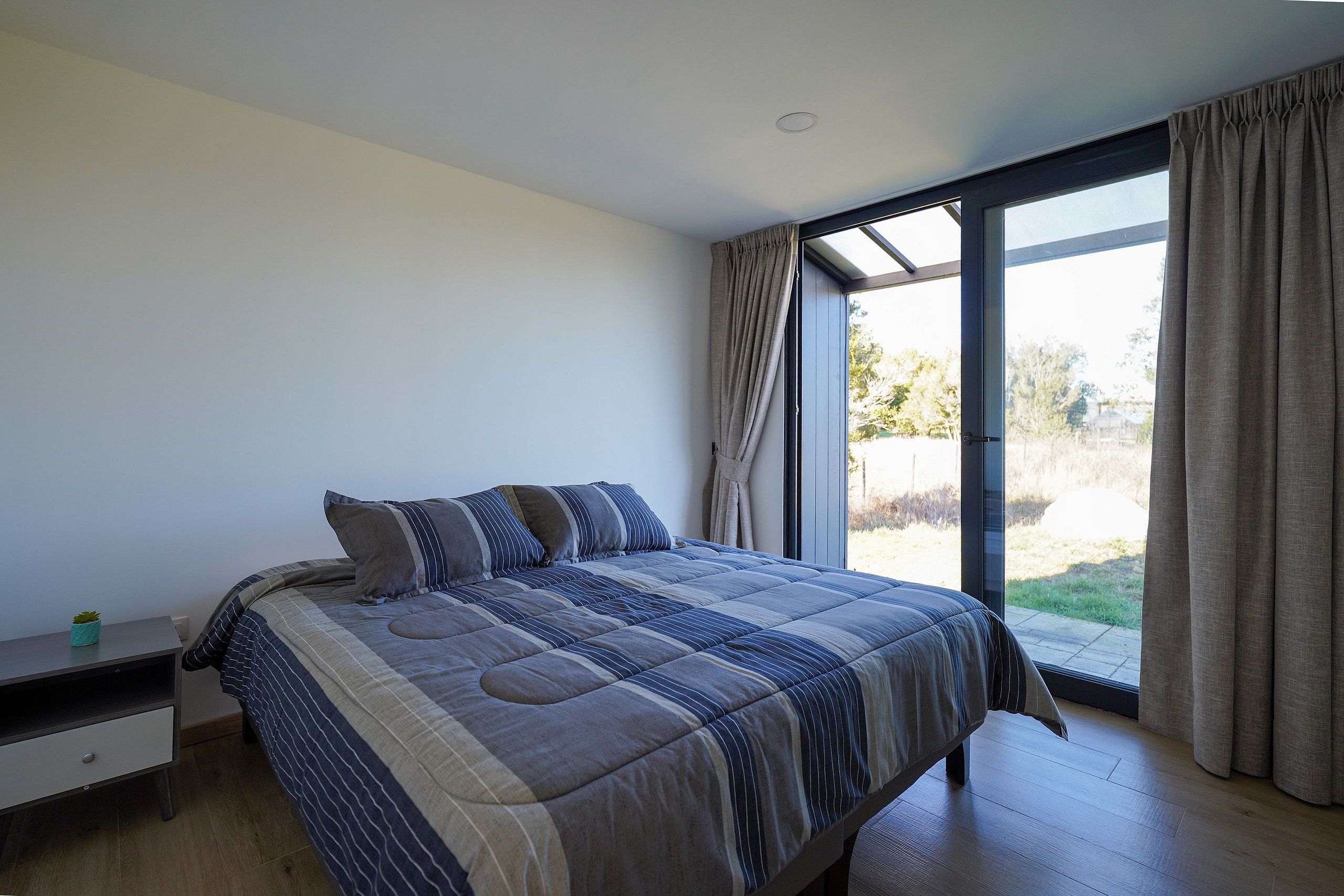 A bedroom with a large bed, a nightstand, a window with curtains, and a view of the outdoors.