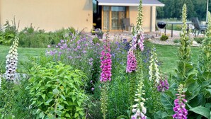 Une maison avec un toit rouge, un jardin où poussent différentes sortes de fleurs et une terrasse aménagée avec du mobilier d’extérieur.