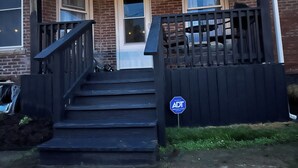 A brick building with a front door, a staircase leading up to the entrance, and a black fence with a sign. - 4 bedroom beautiful modern apartment near Data center fully furnished  (Michigan City)