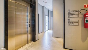 A modern elevator with a metallic finish, a fire extinguisher mounted on the wall, and a corridor leading to a door with a glass panel.