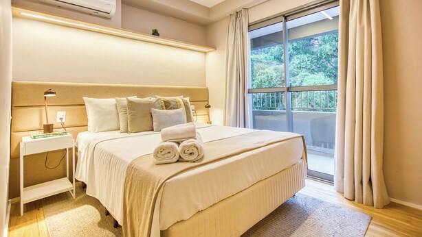 A hotel room with a large bed, a bedside table, an air conditioning unit, and a view of greenery through a window.