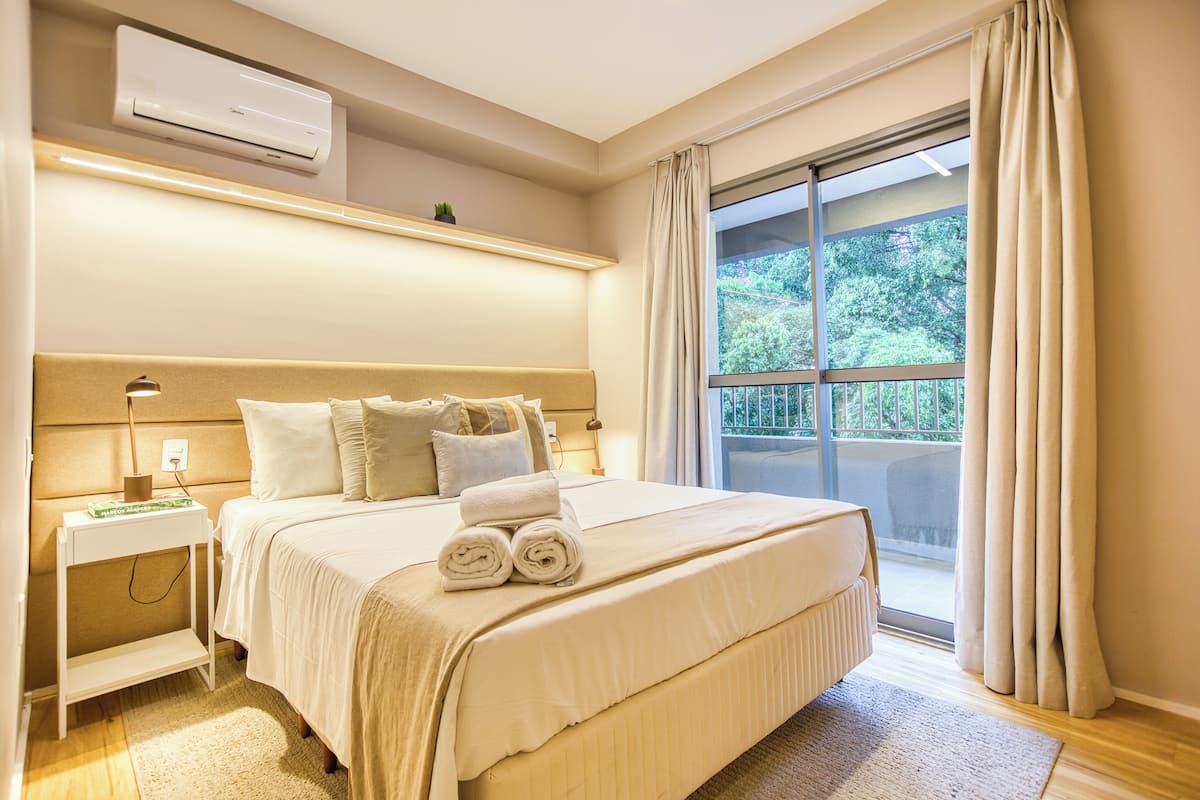 Ein Hotelzimmer mit einem großen Bett, einem Nachttisch, einer Klimaanlage und Blick auf Grün durch ein Fenster.