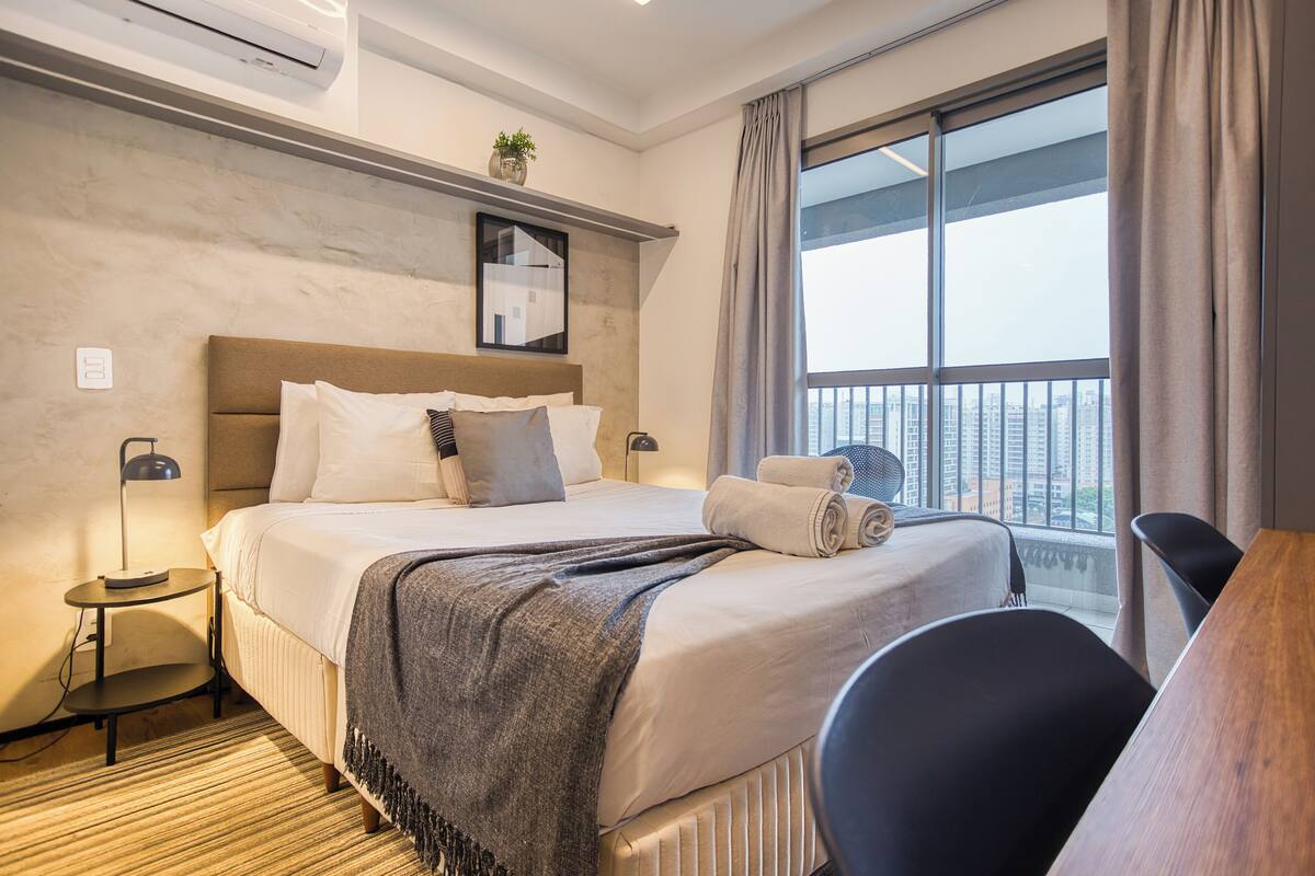 Ein Hotelzimmer mit einem großen Bett, einem Schreibtisch, einem Stuhl, einem Fenster mit Blick auf Gebäude und einem kleinen Tisch mit einer Lampe.