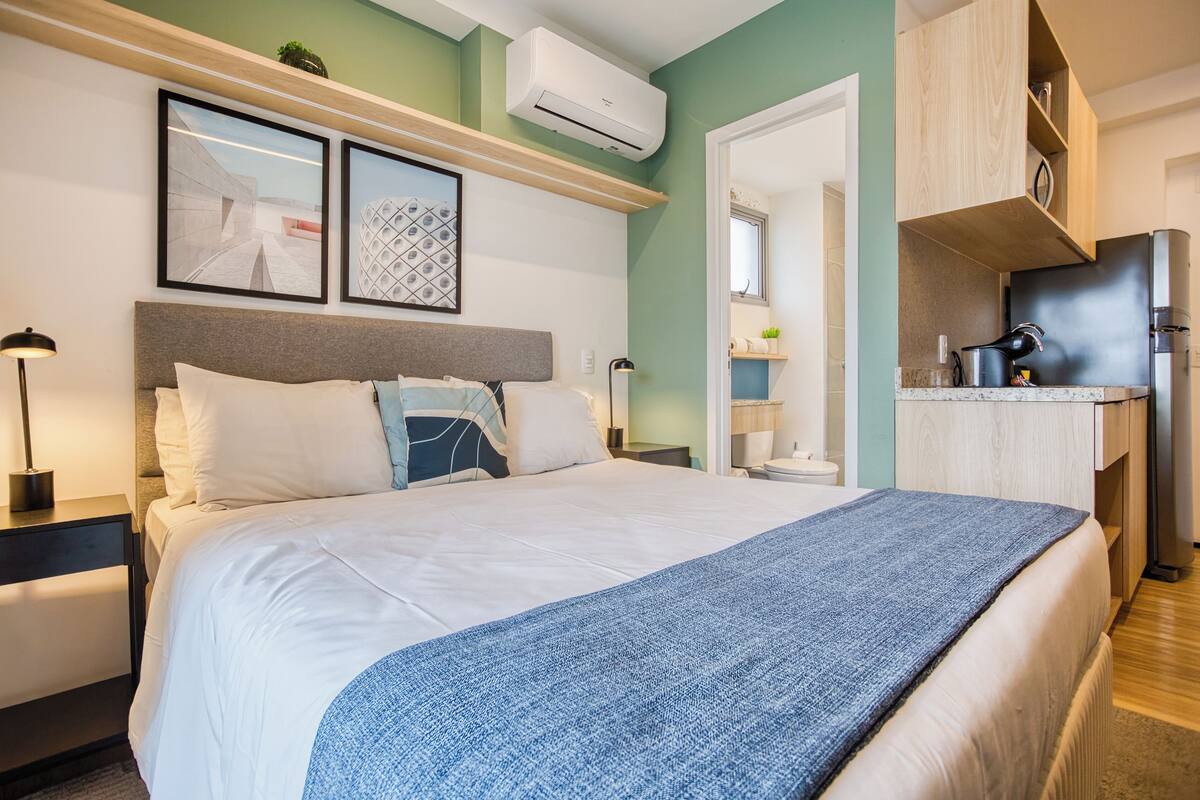 Ein Schlafzimmer mit Bett, Nachttischen, Fenster und einer Küchenzeile mit Kühlschrank und Herd.