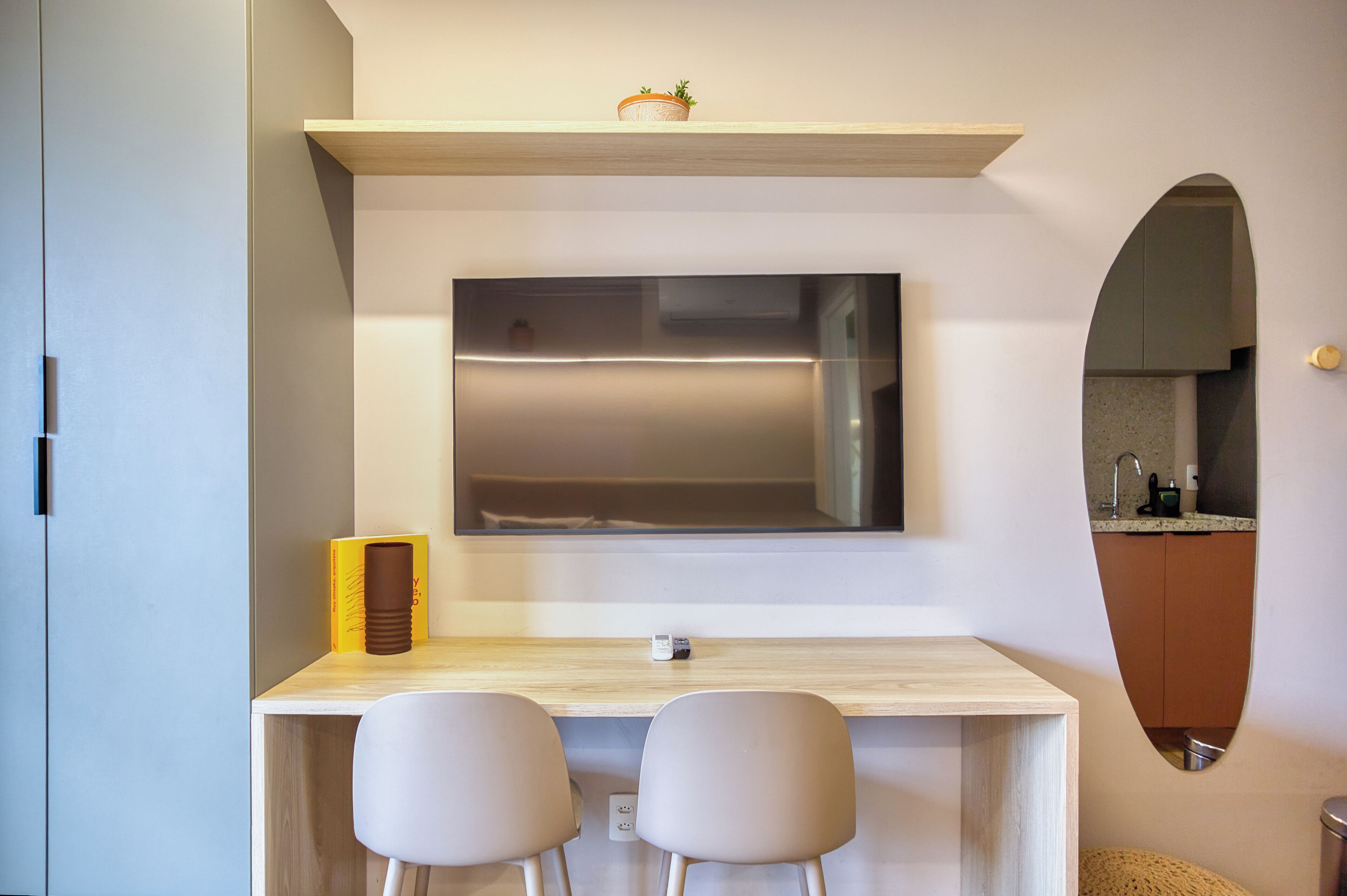 A modern room with a wooden desk, two chairs, a flat-screen TV mounted on the wall, and a small potted plant on a shelf above the desk.