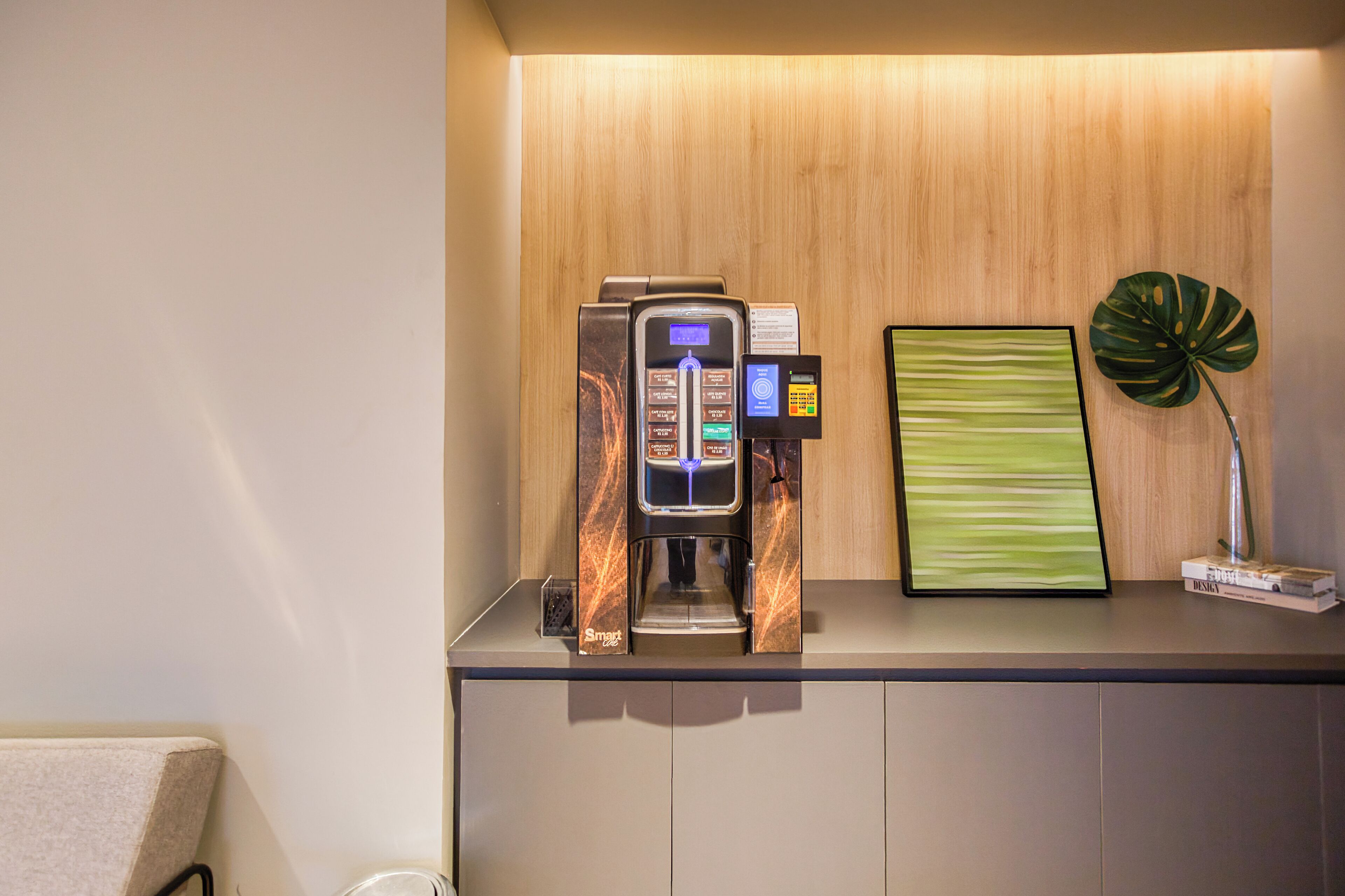 Una máquina de café con pantalla digital y lector de tarjetas, un cuadro abstracto enmarcado y una planta en maceta sobre una encimera.