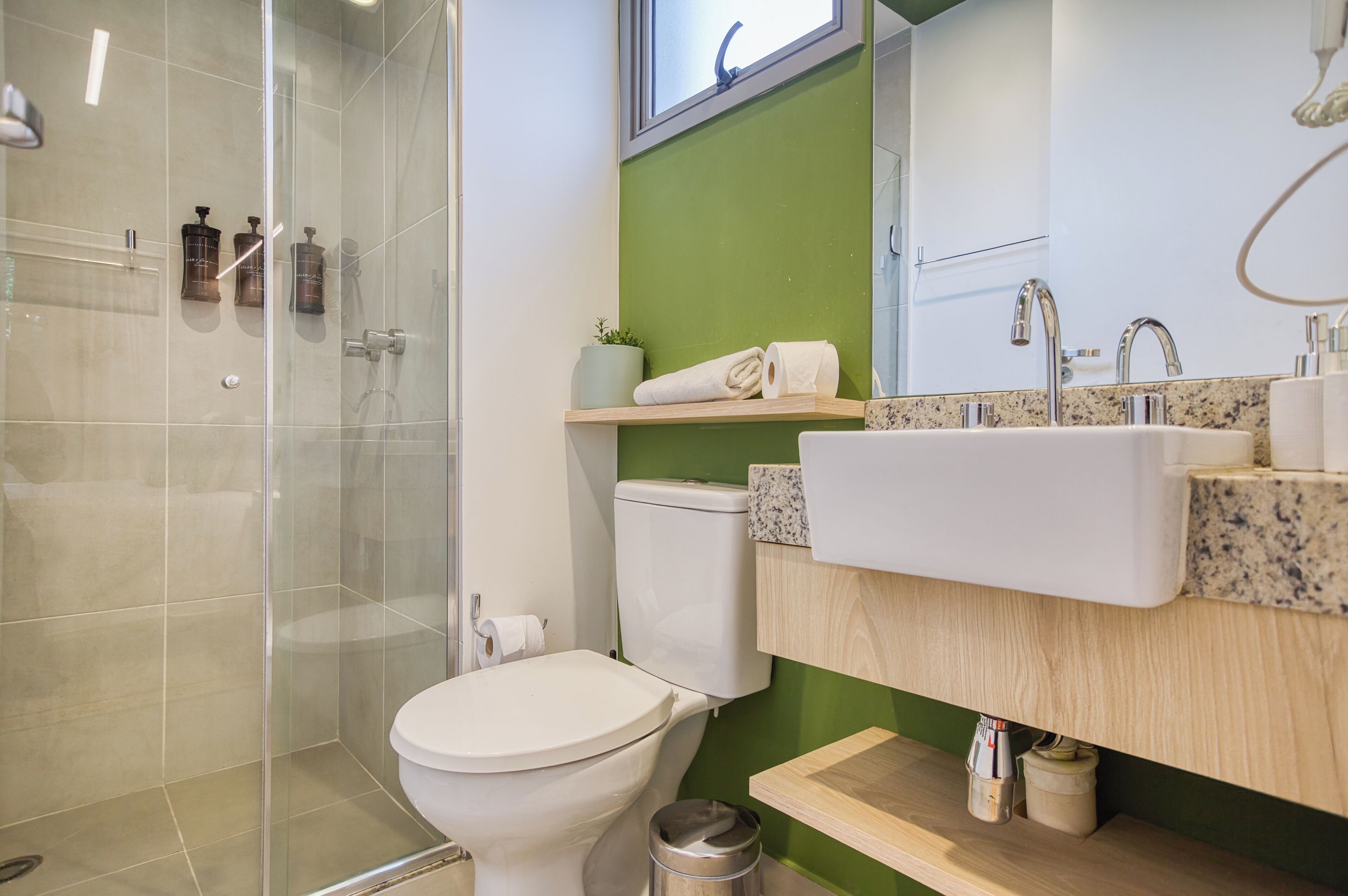 A modern bathroom with a glass shower enclosure, a white toilet, a green accent wall, a wooden vanity with a sink, and a small potted plant.