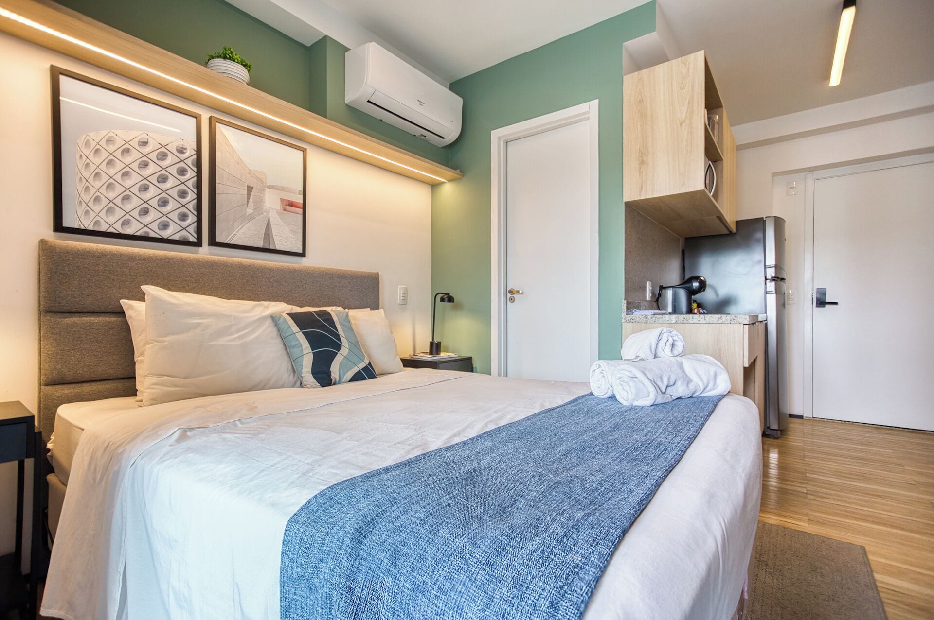 A modern hotel room with a large bed, a bedside table, a nightstand, an air conditioning unit, a framed picture, a towel rack, and a small kitchenette area.