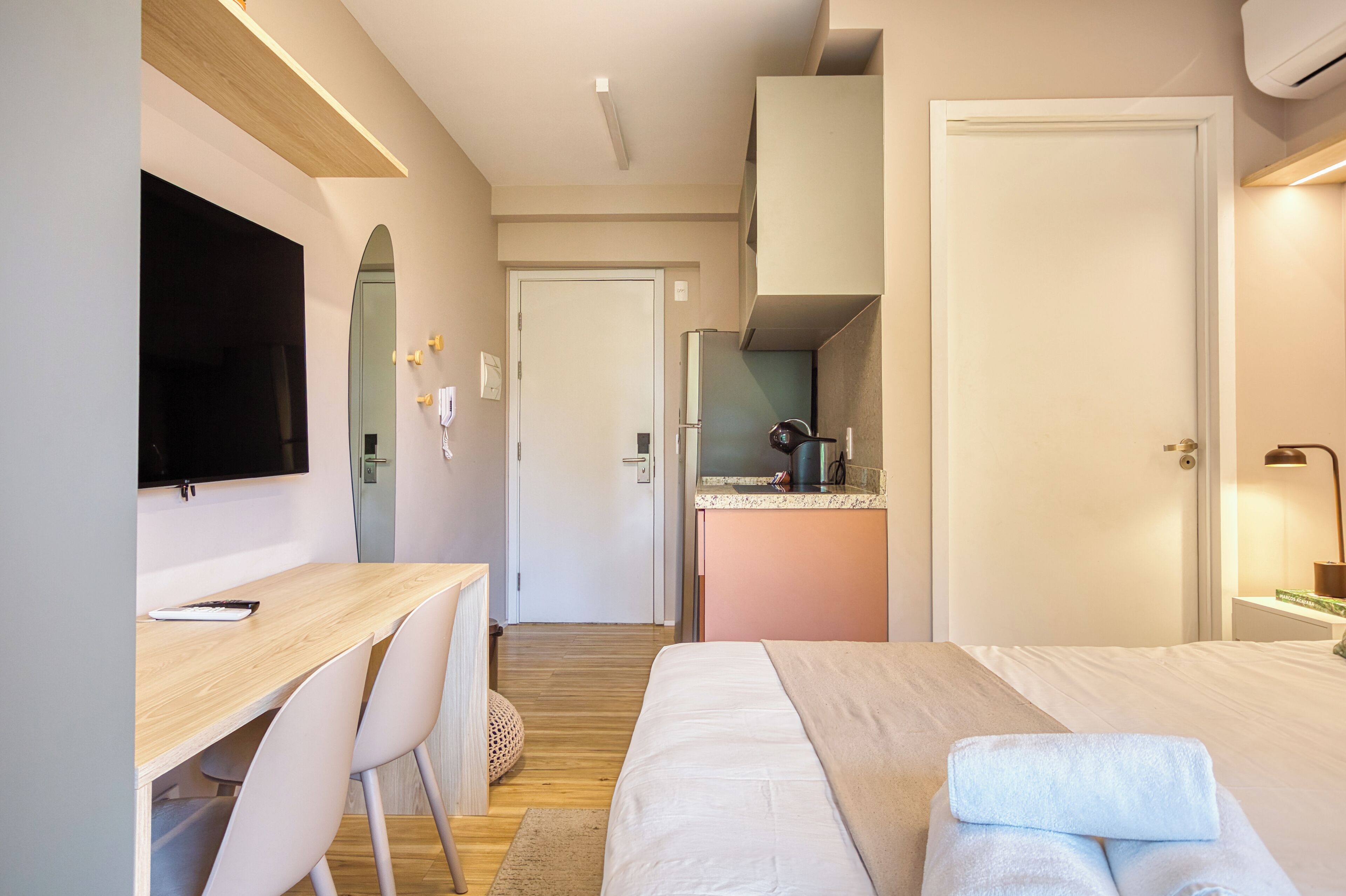A modern hotel room with a wooden desk, a bed with a white comforter, a television, a kitchenette, and a door leading to another room.