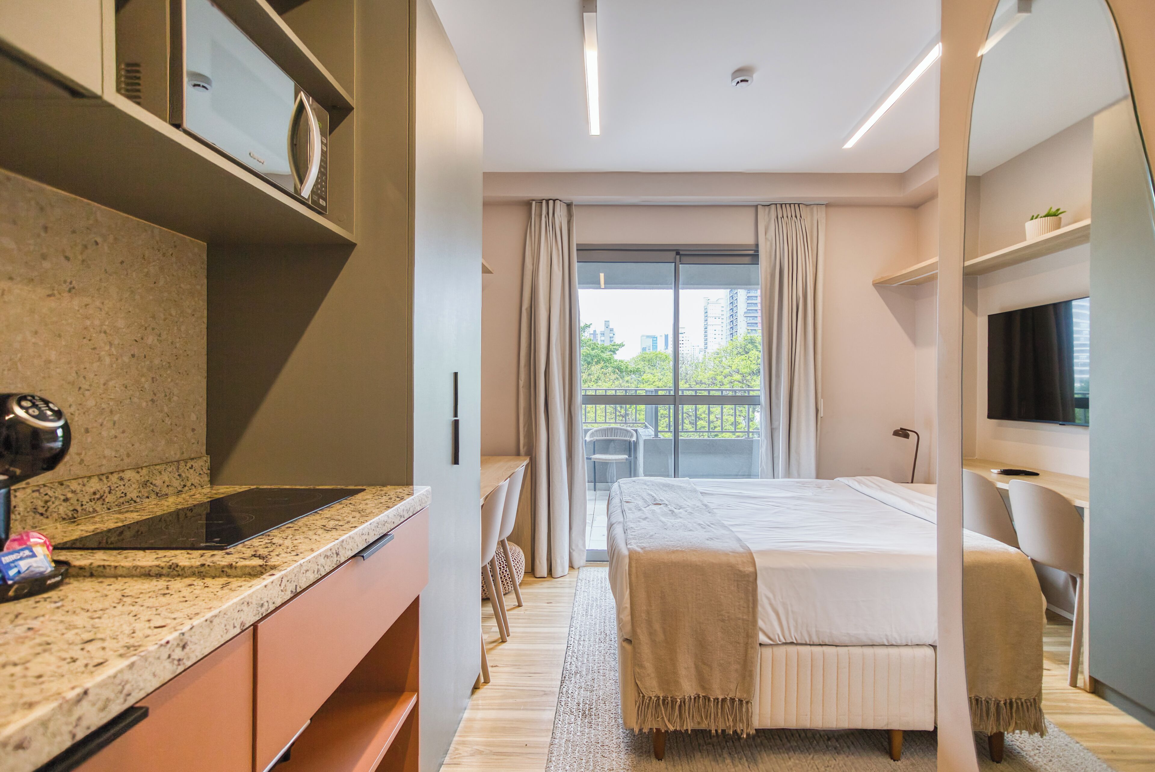 A modern hotel room with a kitchen area, a bed with a view, and a TV.