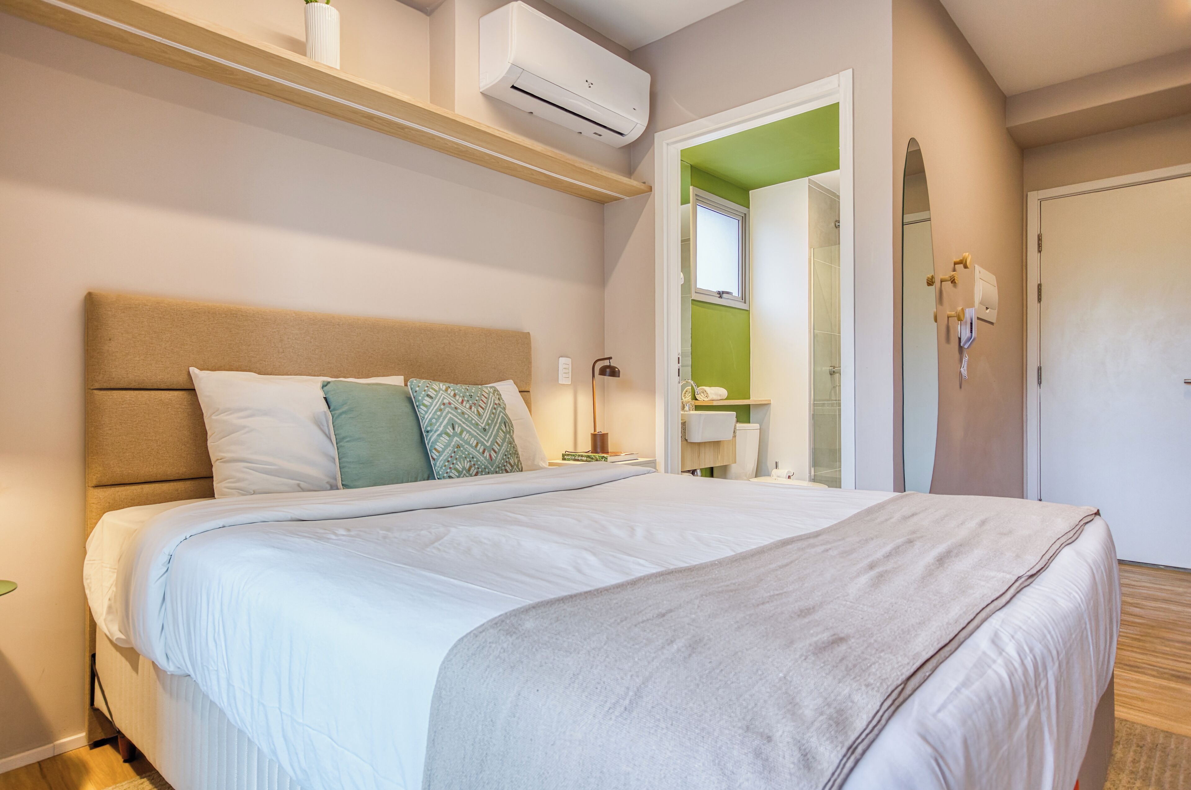 A neatly made bed with a headboard, a bedside table with a lamp, and a wall-mounted air conditioner.