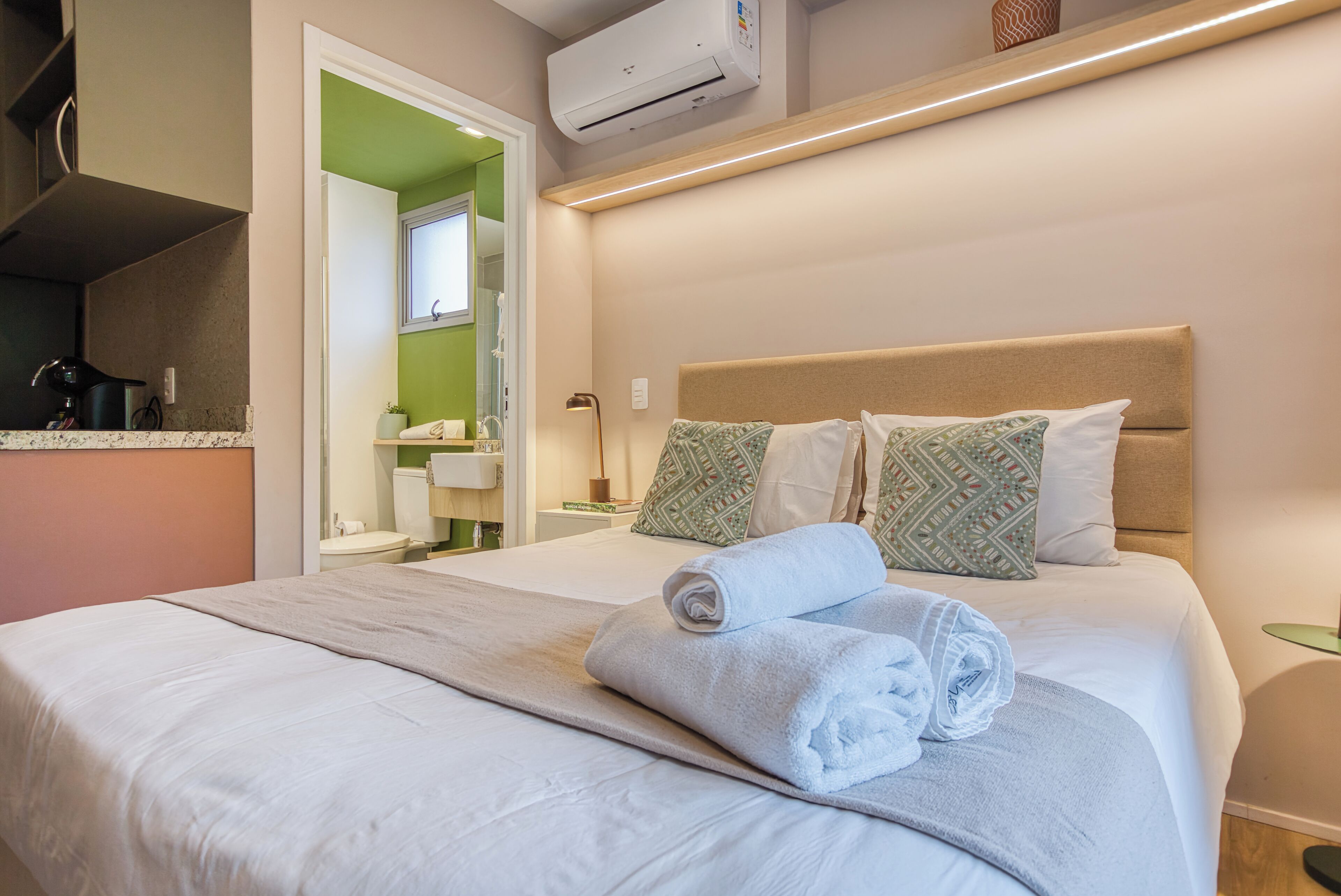 A neatly made bed with a folded towel, a bedside table with a lamp, and a mirror above the sink in a bathroom.