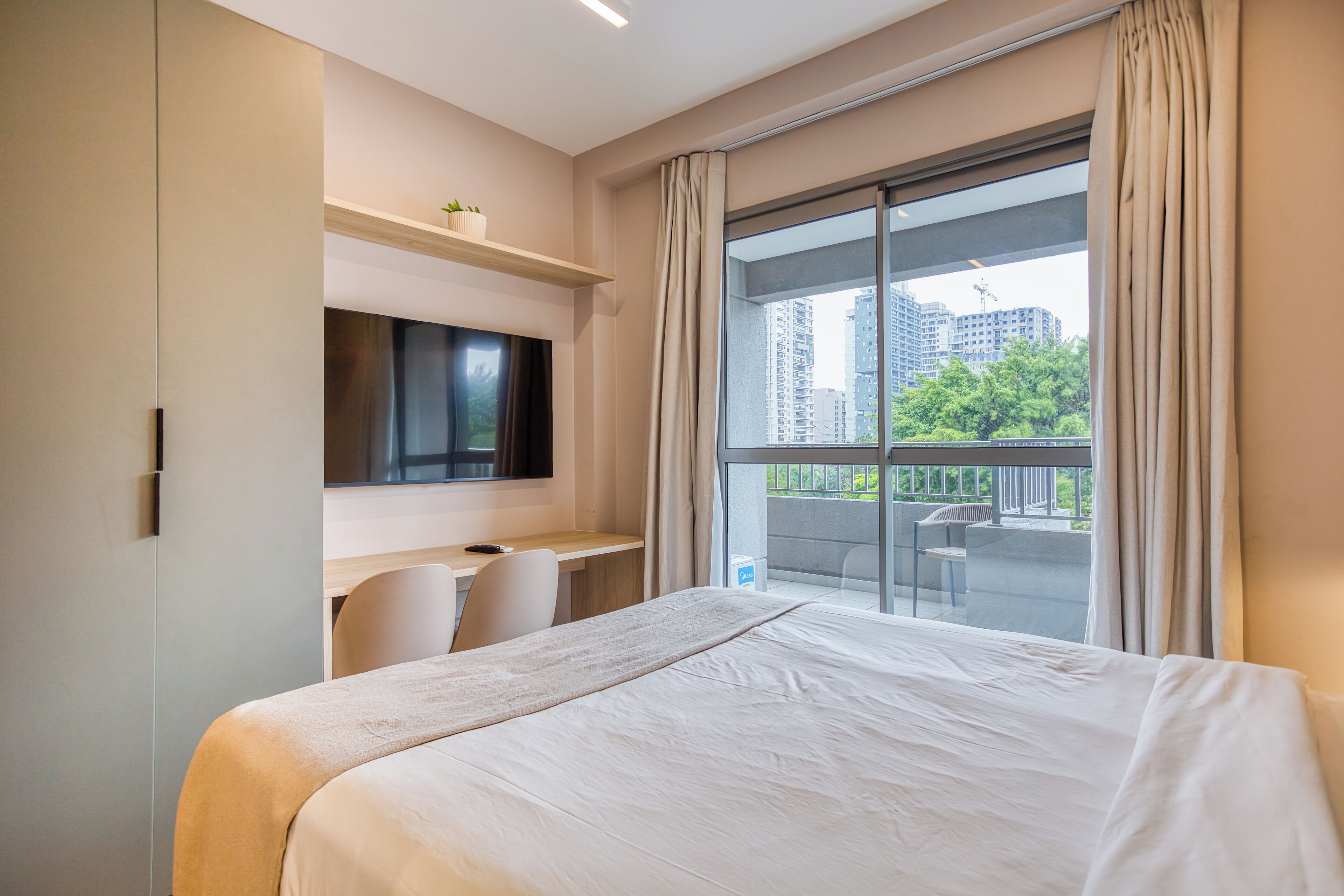 A hotel room with a large bed, a desk with a chair, a TV mounted on the wall, and a balcony with a view of the city.