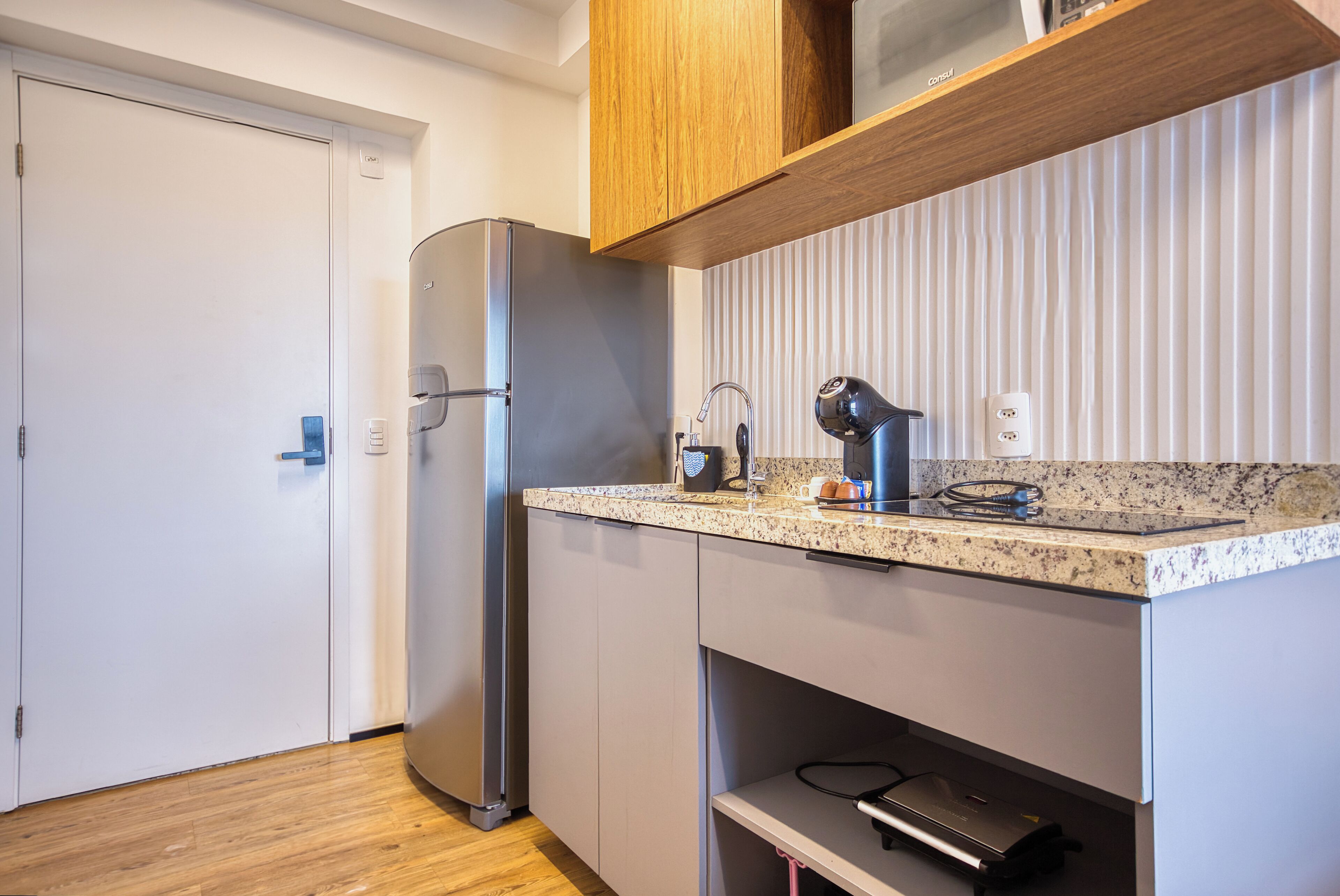 A compact kitchen with a built-in refrigerator, a countertop with a kettle, and a power strip.