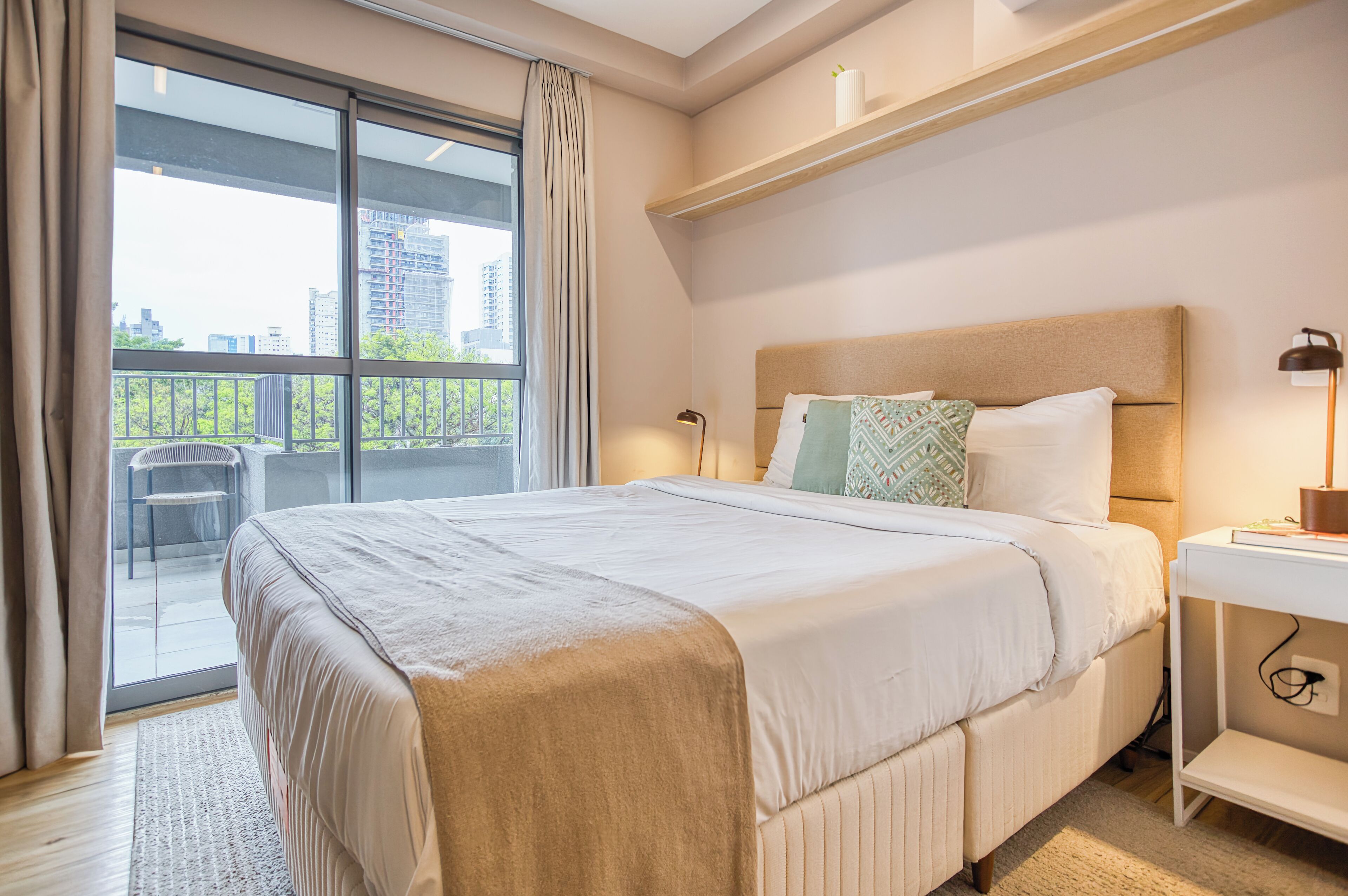 A hotel room with a large bed, a nightstand, a lamp, and a view of the city through a balcony.