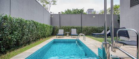 A rectangular swimming pool with a tiled edge, surrounded by a low hedge and a grey wall. There are lounge chairs and a metal ladder leading into the pool.