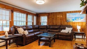 A living room with a sectional sofa, a glass coffee table, wooden flooring, and large windows with blinds. - Naples Village Retreat - Heart of the Lakes Region (Naples)