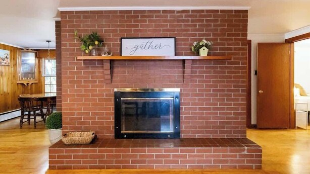 A brick fireplace with a framed sign above it, a small shelf with decorative items, and a basket on the floor. - Naples Village Retreat - Heart of the Lakes Region (Naples)