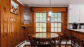 A wooden-paneled dining area with a round table and chairs, a hanging light fixture, and a view through the windows. - Naples Village Retreat - Heart of the Lakes Region (Naples)