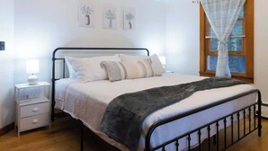A bedroom with a metal bed frame, white bedding, a bedside table with a lamp, and a window with curtains. - Naples Village Retreat - Heart of the Lakes Region (Naples)