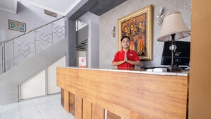A reception desk with a wooden counter, a person standing behind it, a staircase with a geometric railing, and a wall with a framed painting. - Hotel O Near Tugu Yogyakarta 1 formerly Crown Residence (Yogyakarta)