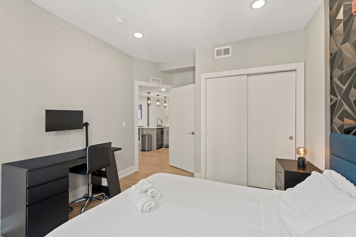 A modern hotel room with a bed, a desk, a chair, a TV, a wardrobe, and a bathroom visible in the background.