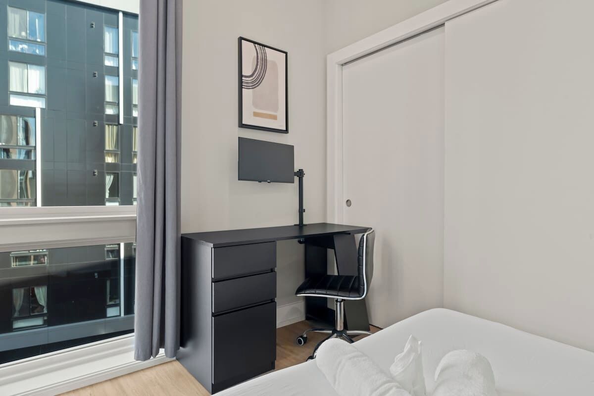 A modern bedroom with a large window, a black desk with drawers, a chair, and a bed with white bedding.