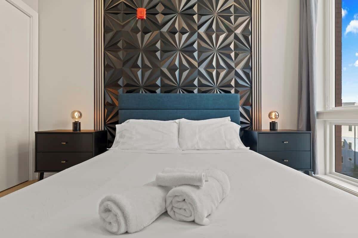 A neatly made bed with white linens and rolled towels, a geometric patterned headboard, and two bedside tables with lamps.