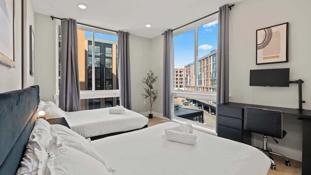 Habitación de hotel con dos camas, un escritorio, una silla, televisión y ventanas amplias con vista a la ciudad.