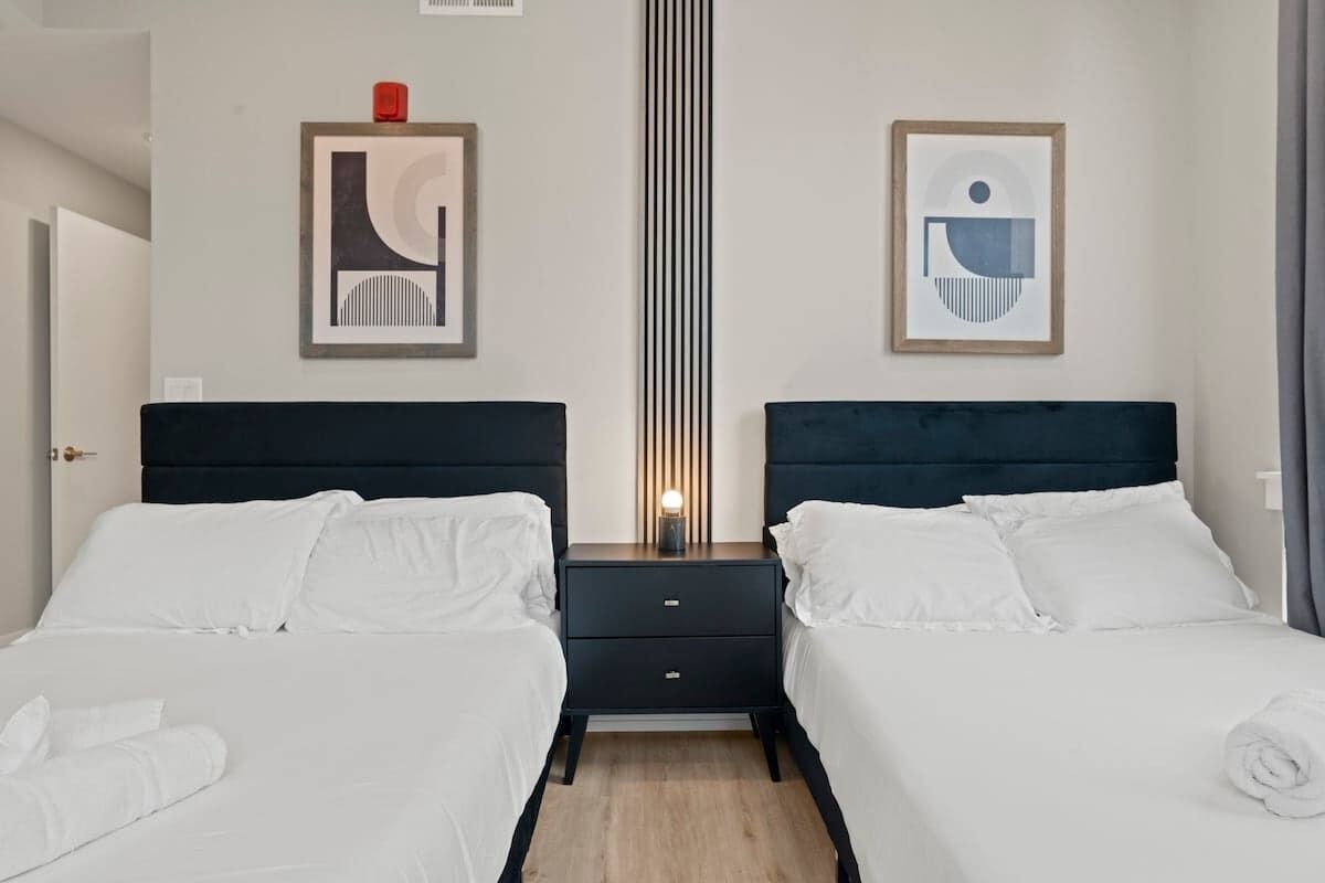 Habitación de hotel con dos camas, una mesita de noche con lámpara y dos obras de arte abstracto enmarcadas en las paredes.