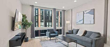 A modern living room with a grey sofa, a coffee table, a TV on a black stand, and a large window with curtains.