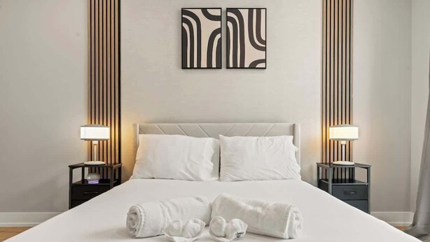 A neatly made bed with white linens and two pillows, flanked by bedside tables with lamps, in a room with vertical paneling on the walls. - Chic 1BR Luxury Apartment in Philly for 4 Guests (Philadelphia)