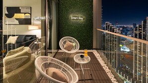 Un balcón moderno con sillas de mimbre, una mesita y barandilla de vidrio con vista a un paisaje urbano nocturno.