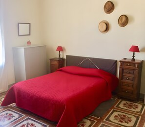 Ein Bett mit roter Decke, zwei Nachttische mit Lampen, eine Kommode und eine Wand mit Hüten.