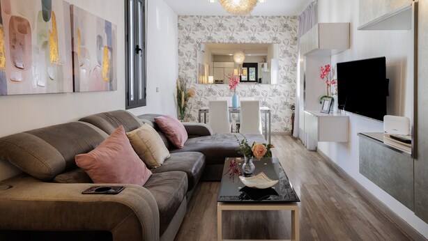 Un salon moderne avec un canapé d’angle, une table basse agrémentée d’objets décoratifs, un téléviseur à écran plat et, au fond, un coin cuisine.