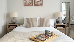 Quarto bem arrumado com uma cama, duas mesas de cabeceira, um criado-mudo, um tabuleiro com livros e canecas, e duas fotografias emolduradas na parede.
