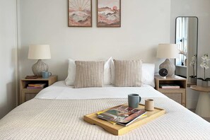 Ein ordentlich eingerichtetes Schlafzimmer mit einem Bett, zwei Nachttischen, einem Beistelltisch, einer Ablage mit Büchern und Tassen sowie zwei gerahmten Bildern an der Wand.