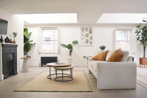 A modern living room with a sofa, a coffee table, and a fireplace. There are several potted plants and a large window providing natural light.