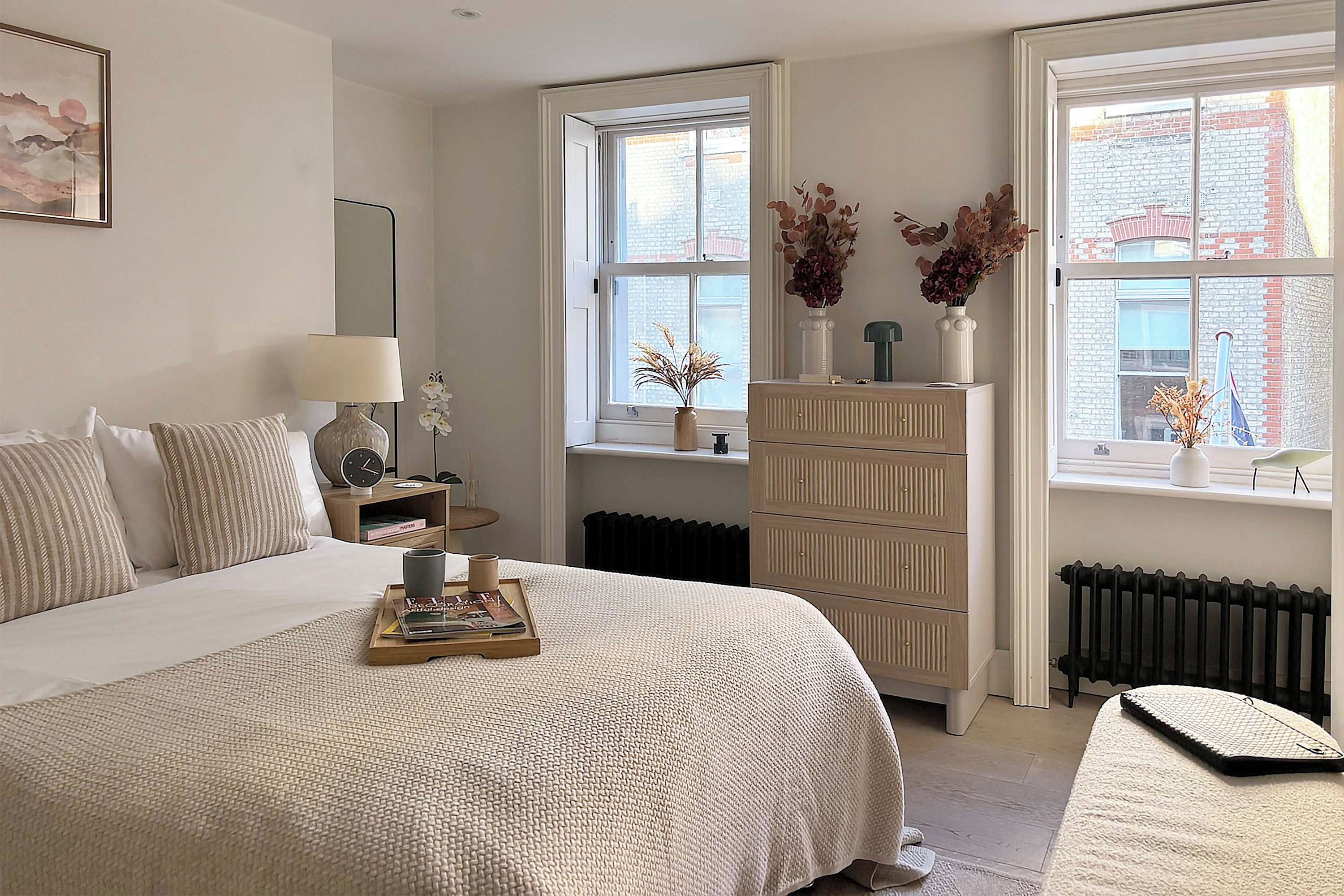Une chambre à coucher comprenant un lit, une table de chevet, une lampe, une commode et une fenêtre donnant sur des immeubles.