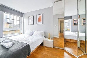 A modern bedroom with a large bed, a bedside table, a mirror, and a window with a city view. - Canary Wharf Skyline Views (Greater London)
