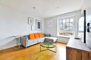 A modern living room with a grey sofa, orange cushions, a glass coffee table, and a large window with a city view. - Canary Wharf Skyline Views (Greater London)