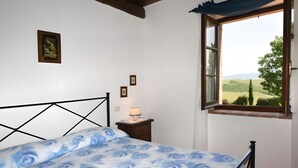 A bedroom with a bed, a nightstand, a lamp, a window with a view of the countryside, and a framed picture on the wall. - Apartment 'Azzurro La Valle Del Sole' with Mountain View and Wi-Fi (Castiglione dʼOrcia)