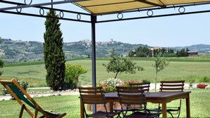 Un espacio de estar techado al aire libre con una mesa y sillas, con vistas a un paisaje verde con árboles y edificios al fondo.