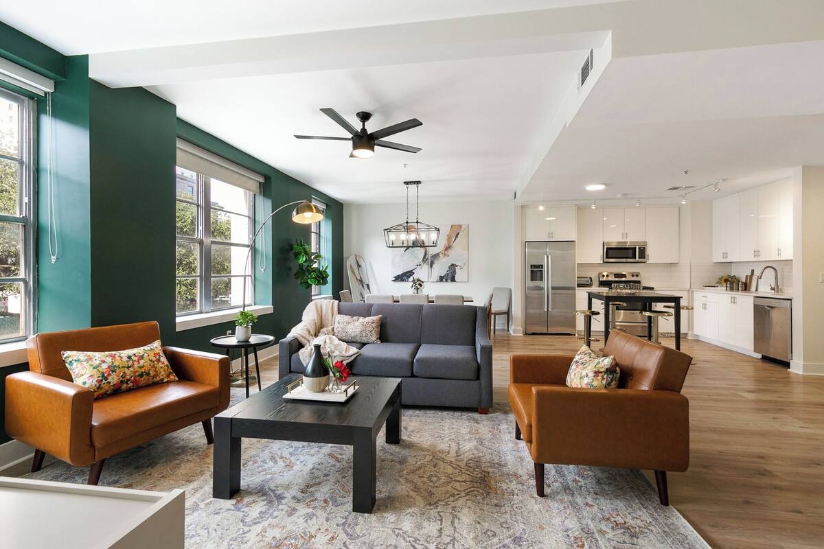 A spacious living room with a grey sofa, brown leather armchairs, and a coffee table. A dining area with a chandelier and a kitchen with white cabinets and stainless steel appliances are visible in the background.