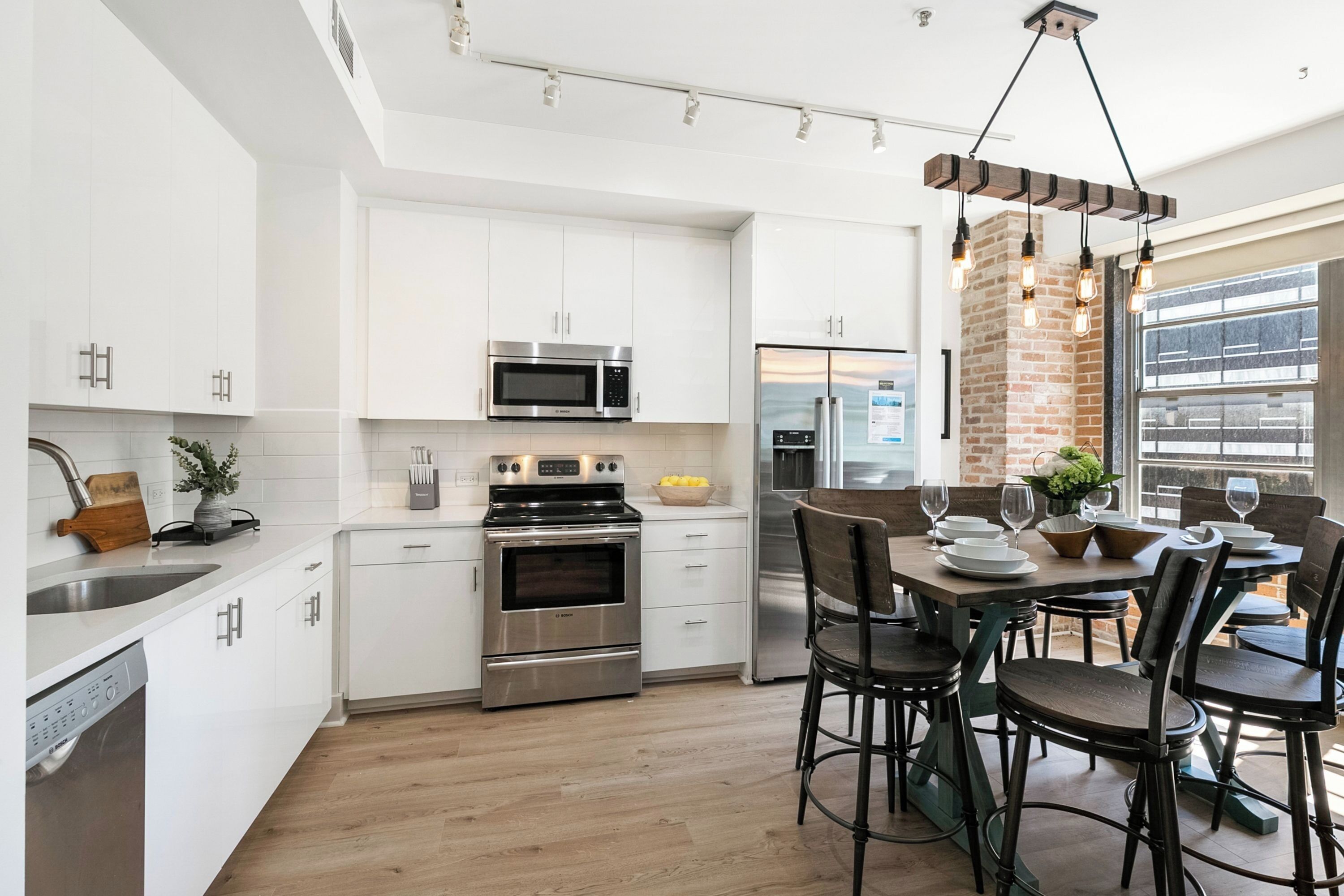 Una cocina moderna con gabinetes blancos, electrodomésticos de acero inoxidable y una zona de comedor con pared de ladrillo y lámparas colgantes.