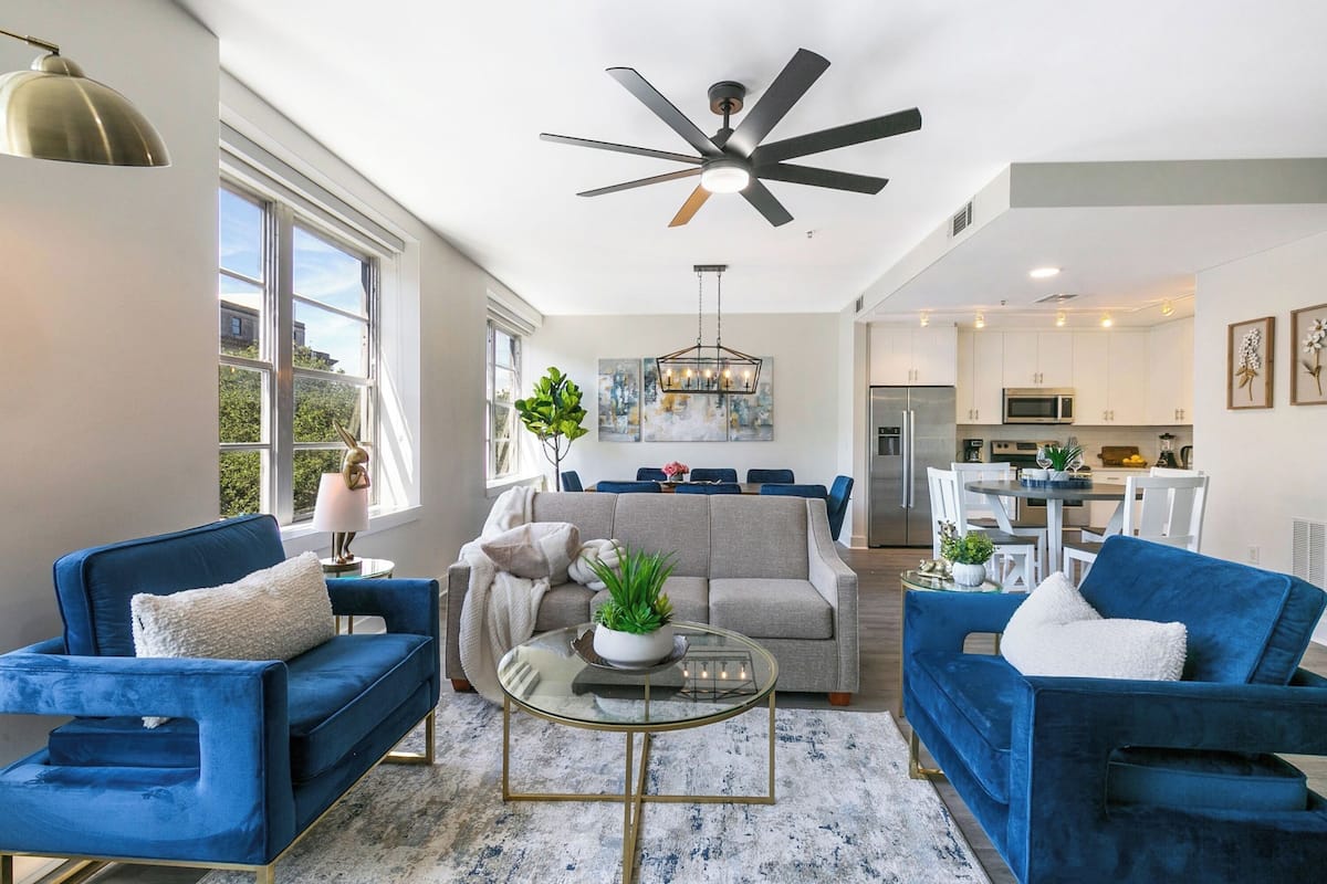 A modern living room with blue sofas, a glass coffee table, and a ceiling fan.